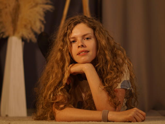 Tamekia Iafrate, een blanke vrouw met lang, krullend bruin haar, leunt op haar ellebogen en kijkt verleidelijk. Het warme licht accentueert haar gezicht en haar, waardoor een sensuele, intieme sfeer ontstaat. Zachte achtergrond..