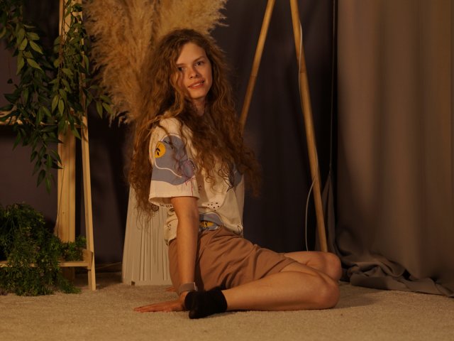 Tamekia Iafrate zit op een tapijtvloer, met gebogen benen, en draagt een witte blouse met blauwe patronen en bruine korte broek. Ze heeft lang, krullend haar, ze glimlacht, en er staat een plant en een houten standaard achter haar. Heel aantrekkelijk..