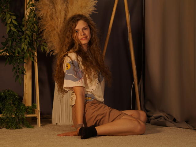Tamekia Iafrate est assise par terre, sur un tapis, les jambes pliées, portant une chemise blanche avec des motifs colorés et un short marron, long. Cheveux bouclés, chaussettes foncées, appuyé contre une plante en pot, souriant. Éclairage chaleureux.