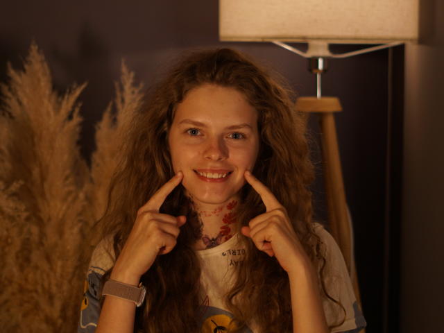 Foto van Tamekia Iafrate, een jonge vrouw met lang, krullend bruin haar, die glimlacht en met haar vingers naar haar wangen wijst. Ze draagt een witte blouse, een kralenketting en een armband, in warm, binnen licht..