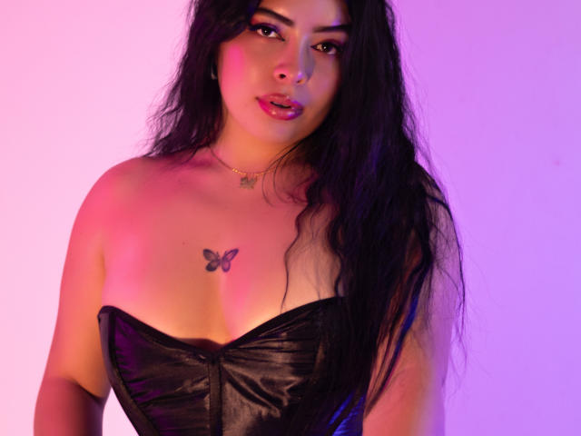 Kate Welsin, com longos cabelos pretos, vestindo uma blusa de couro preta sem alças, iluminação em tons de roxo e rosa, tatuagem de borboleta no peito, olhando diretamente para a câmera com os lábios ligeiramente entreabertos. Muito quente.