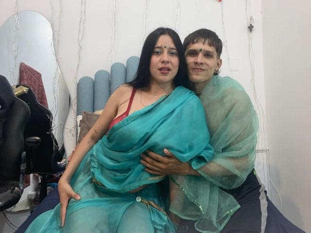 AnjaliRohan, a brown-skinned woman with black hair, wearing a turquoise saree, sits with her back to a brown-haired man in a translucent green shawl. He's grabbing her waist, erection visible.