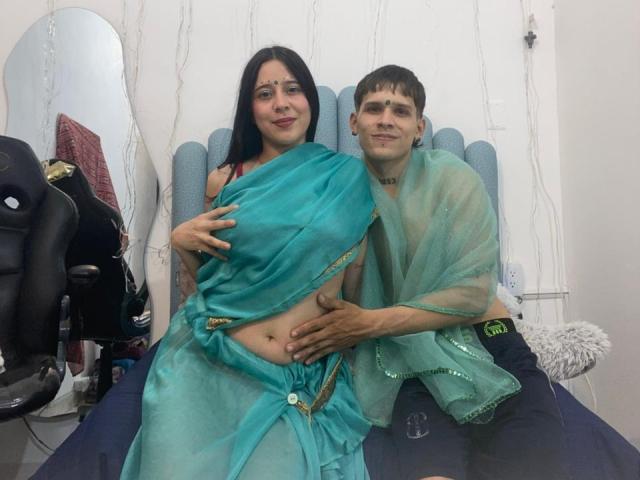 AnjaliRohan with dark hair and green saree, sitting on a chair, wearing a translucent green shawl, with a slim, fair-skinned man in black pants, hand on her stomach. Sexual tension.
