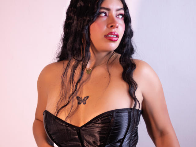 Foto van KateWelsin met lang zwart haar, die een zwarte bustier zonder bandjes draagt, met een tatoeage van een vlinder op haar lichaam. borst. Ze heeft rode lippenstift en een verleidelijke uitdrukking, en kijkt omhoog..