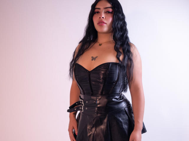 Kate Welsin, com longos cabelos pretos, vestindo um corpete preto justo e uma saia de couro, em pé contra um fundo branco. Tatuagem no peito, quadris arqueados, olhar intenso, tensão sexual, expressão sedutora..