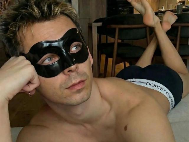 Photographie de dannyboy20-fun-hot, allongé sur un lit, portant un masque noir et un sous-vêtement noir avec une bande de taille blanche, jambes. Accroupi, les genoux pliés, la tête reposant sur sa main. Cocorico visible.