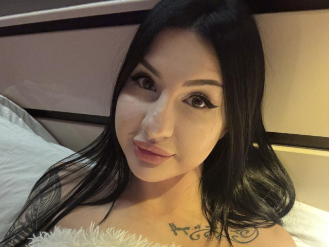 Close-up selfie van AuraXo, een Aziatische vrouw met lang zwart haar, dikke eyeliner en een subtiele glimlach. Ze draagt een pluizig wit topje, waar een kleine tatoeage van een vogel op haar borst te zien is. Haar blik is direct en straalt een subtiele seksuele spanning uit..