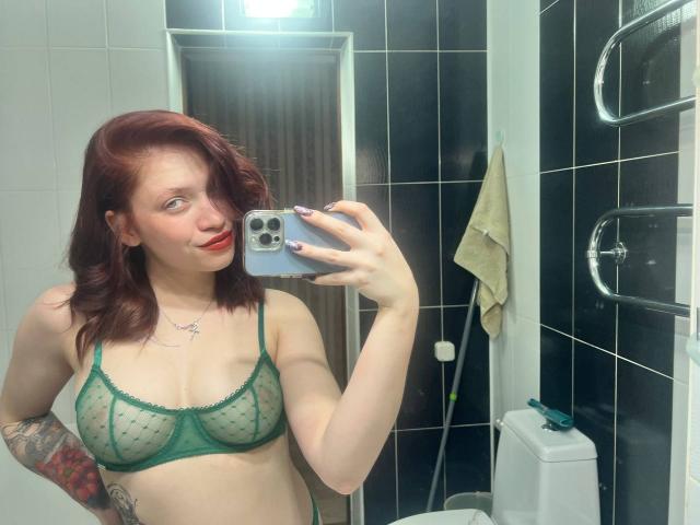 Peggy Behimer, met haar lichte huid, rode haar en tatoeages, maakt een foto van zichzelf in een zwart betegelde badkamer. Ze draagt een groene, doorschijnende BH, ze glimlacht en houdt een telefoon met een zilveren hoesje vast..