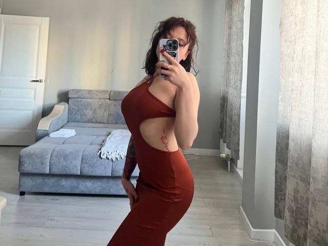 PeggyBehimer se toma un selfie frente a un espejo en una sala de estar, vistiendo un vestido rojo ajustado con un solo hombro y un corte, mostrando su senos grandes y caderas curvilíneas. Ella está sosteniendo un teléfono con una funda estampada..