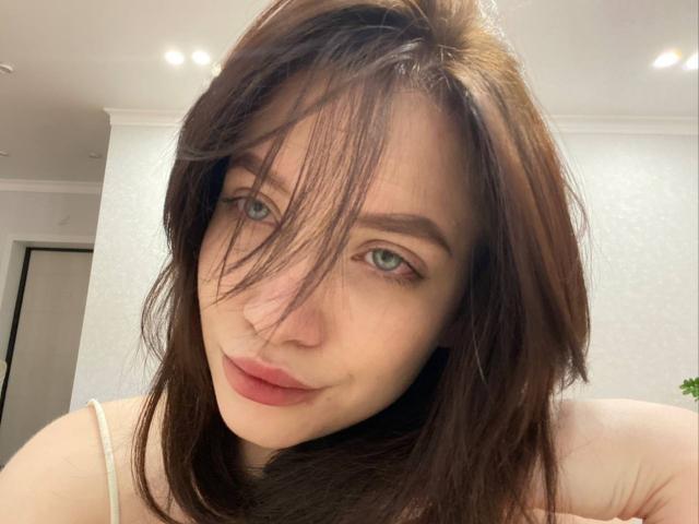 Primer plano de PeggyBehimer, una joven con piel pálida, ojos verdes y cabello castaño oscuro, mirando directamente a la cámara. con una leve sonrisa. El cabello cubre parcialmente su rostro. Iluminación brillante..