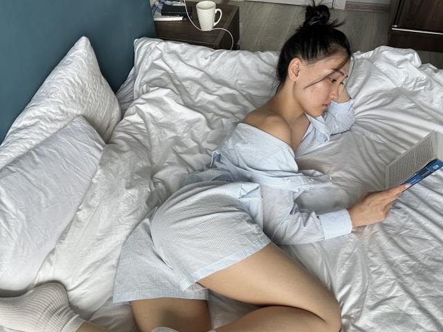 EvaLena mit dunklem Haar zu einem unordentlichen Dutt gebunden, trägt ein weißes Top mit einem Schulterauschnitt und liegt in einem unaufgeräumten Bett, während sie ein Buch liest. ihr nacktes Schulterblatt, sie sah verführerisch und entspannt aus..