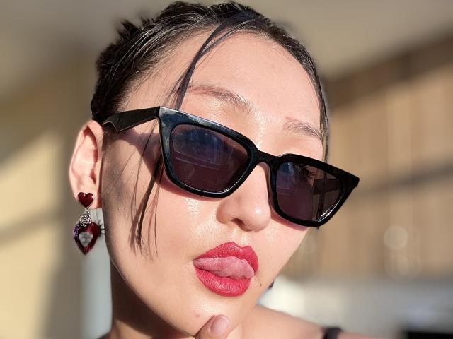 Fotografía de primer plano de EvaLena con gafas de sol negras, lápiz labial rojo y cabello oscuro recogido en un moño. Ella lleva aretes en forma de corazón rojo. La luz del sol brillante resalta su rostro. Increíblemente atractiva, con una vibra segura..