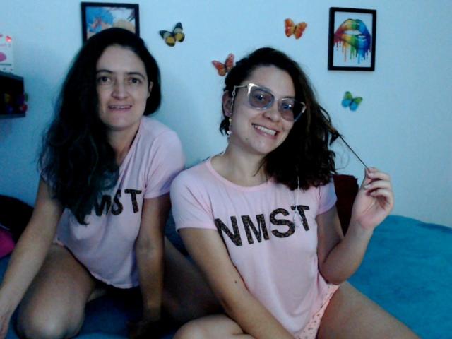Dos mujeres con cabello negro rizado, que llevan camisetas rosas con la inscripción "NMST" y gafas, sentadas en una cama azul. Una persona sostiene el cabello, ambos sonriendo. Decoraciones de pared con forma de mariposa y pinturas coloridas en el fondo..