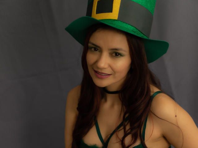 Fotografia di KateBurning, una donna latina con lunghi capelli scuri, che indossa un cappello a cilindro verde con una fibbia gialla e un collare, sorridente con rossetto scuro. Ha un'aria seducente..