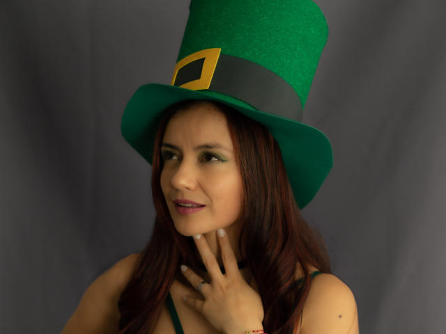 KateBurning indossa un alto cappello verde con una fascia nera e una fibbia dorata, guardando seducentemente la fotocamera. con un dito sul mento. I suoi capelli scuri incorniciano il suo viso..