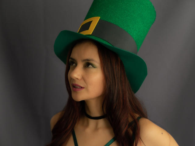 Foto van KateBurning, een aantrekkelijke vrouw met lang bruin haar, die een groene, glinsterende hoed met een zwarte band draagt en gouden gesp, verleidelijk kijkend naar de camera, donkergrijze achtergrond..