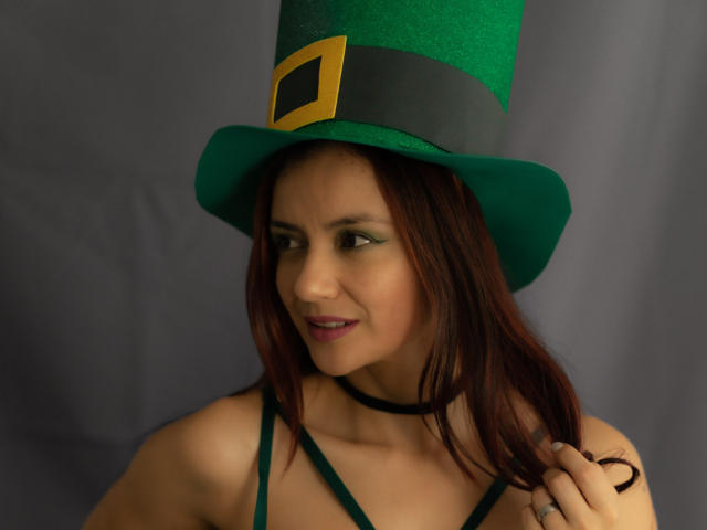 KateBurning, wearing a green top hat with a black band and gold buckle, has dark red hair, black choker, and dark lipstick. She gazes sideways, exuding sexual tension. Photograph.