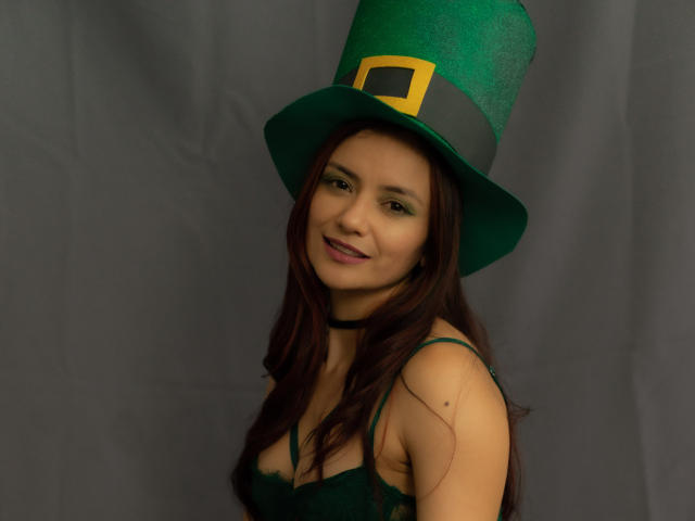KateBurning wears a glittery green top hat with a yellow buckle, smiling with dark green eye makeup. She has long brown hair, black lingerie, and a confident, sexy vibe.