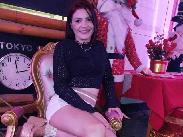 KatteRouse está sentada em uma cadeira de ouro ornamentada, vestindo uma blusa preta de manga comprida e calcinhas brancas, sorrindo. Uma figura de Papai Noel vestida de vermelho está atrás dela, um relógio à esquerda e uma toalha de mesa vermelha..