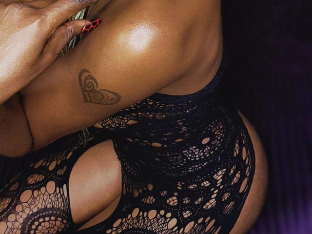 Close-up of FrostyTwinkle's dark-skinned, curvy body in black lace lingerie, showing a butterfly tattoo on the upper arm and red manicured nails. Sexual tension with visible butt and tight fabric.