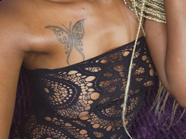Close-up van FrostyTwinkle's schouder en bovenste borst, waarop een zwarte vlindertatoeage te zien is. FrostyTwinkle draagt een zwarte kanten jurk met grote, ronde uitsparingen, en gouden, gevlochten sieraden. Seksuele spanning.