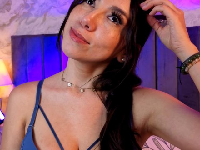 Fotografía de Mia Reed, una mujer de pelo oscuro y largo, que lleva un top azul con tirantes finos, con una mano en el pelo, con una expresión seductora. sonrisa, rostro con pecas, labios rosados, collar con forma de corazón, iluminación azul y morada..