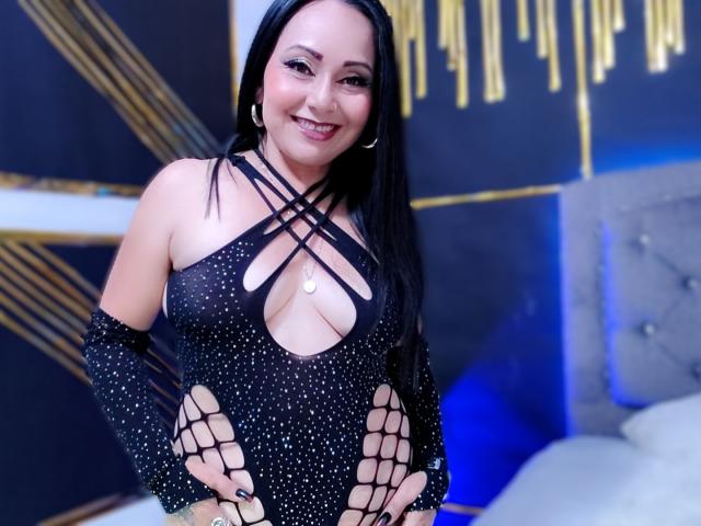 Zaria69 with long black hair, smiling, wearing a black, sparkly, fishnet bodysuit with criss-cross straps and large cutout revealing cleavage. Blue and gold background. Sexy, confident pose.