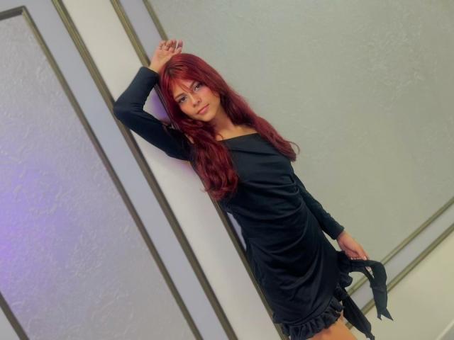 ViioletteRoselle está de pie frente a una pared blanca, apoyada con una mano, vestida con un vestido negro ajustado con bordes deshilachados. Sosteniendo una correa negra con la otra mano. Cabello rojo, mirada seductora.
