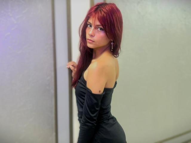 Fotografie von Viiolette Roselle, einer attraktiven jungen Frau mit langen roten Haaren, die ein eng anliegendes, schwarzes Kleid mit einem frechen Ausschnitt trägt. stehend in der Nähe einer vertikalen, weißen Lichtleiste, mit einem verführerischen Blick zurückblickend..