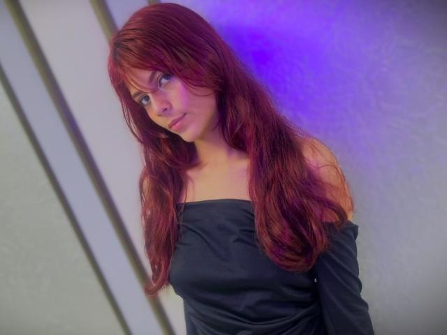 ViioletteRoselle, mit langen roten Haaren und Pony, trägt ein schwarzes Top mit einem Schulter frei, und blickt verführerisch zur Seite. Blaues und violettes Licht betonen sie und erzeugen eine sinnliche, erotische Atmosphäre..