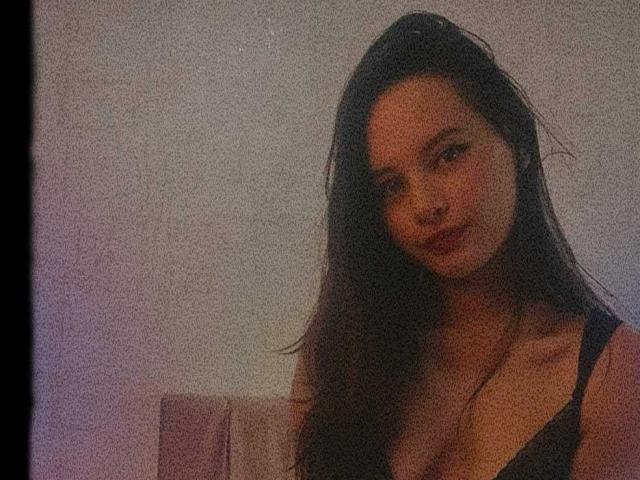 Foto sgranata di LunaBellee, una donna attraente con lunghi capelli scuri, che indossa una canotta nera e si sporge in avanti con un'aria seducente. espressione.
l'illuminazione in tonalità viola e rosa tenue crea un'atmosfera seducente..