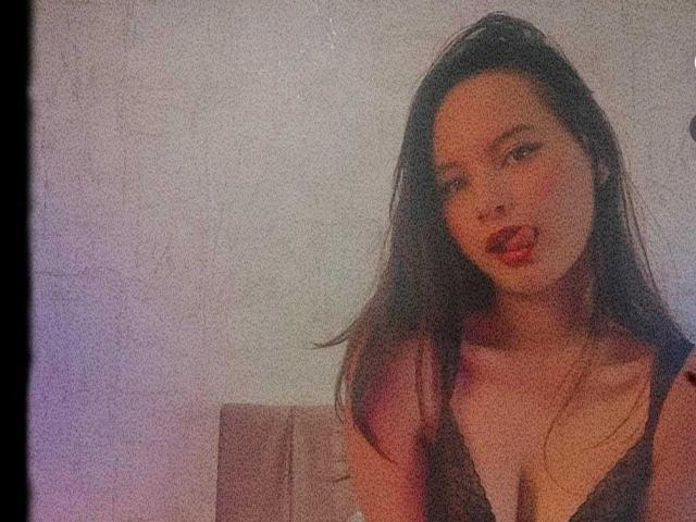 Foto van LunaBellee, een aantrekkelijke Aziatische vrouw met lang zwart haar, rode lippenstift en een diep decolleté, gekleed in een zwarte tanktop. Boven. Haar uitdrukking is verleidelijk, met een lichte kanteling van haar hoofd..