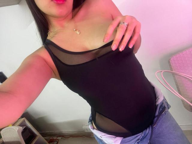 Close-up photo of IrisBlair with her head slightly out of frame, wearing a black sheer top, exposing a hint of her breast. She's touching her collarbone, blue jeans slightly unbuttoned. Pink lighting.