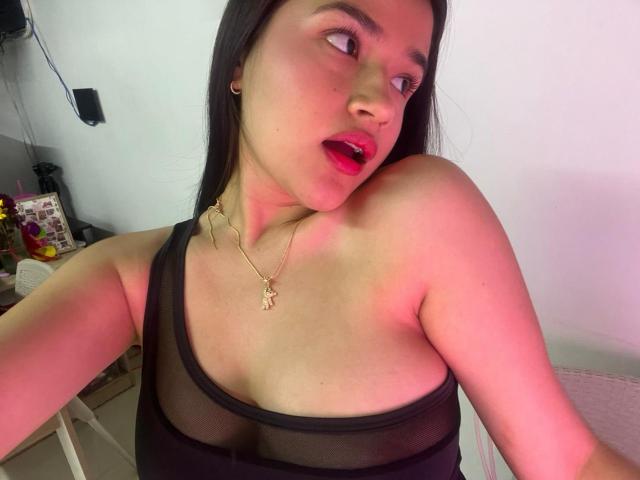 Close-up photo of IrisBlair, an Asian woman with long black hair, wearing a sheer black top that reveals her small breasts. She's looking to the side with red lips, wearing a gold necklace. Pink lighting casts a glow on her skin. She's leaning forward, creating a provocative pose.
