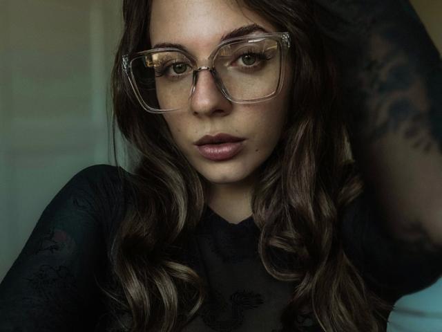 Close-up foto van Junaxwild-hot, een aantrekkelijke vrouw met lang, golvend bruin haar, die een bril met een transparante montuur en een zwarte top, die recht in de camera kijkt met intense, verleidelijke ogen.