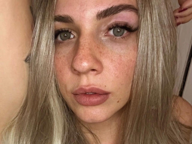 Gros plan de Junaxwild-hot, une jeune femme aux cheveux blonds, aux yeux bleus et aux taches de rousseur. Elle a les lèvres pulpeuses, des sourcils épais et un regard intense. Son expression est sérieuse, avec une légère tension sexuelle..