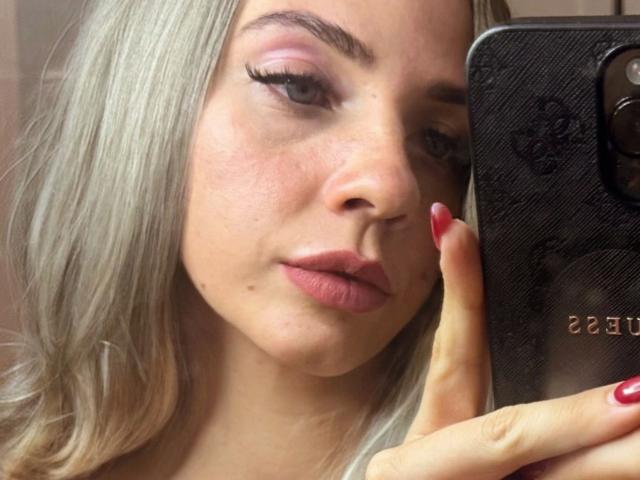 Nahaufnahme eines Fotos von Junaxwild-hot mit heller Haut, platinblondem Haar und roten Nägeln, die ein schwarzes Smartphone hält. Sie blickt mit leicht geöffneten, rosafarbenen Lippen auf das Telefon, was eine sexuelle Spannung erzeugt..