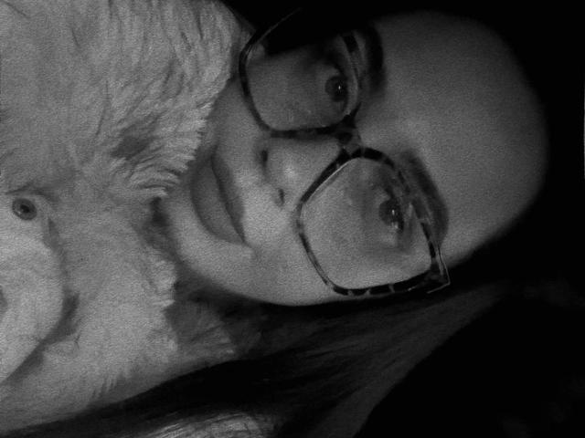 Photographie en noir et blanc, en gros plan, de Junaxwild-hot, portant de grandes lunettes et regardant directement l'appareil photo. Elle a un col en fourrure duveteuse, des cheveux foncés et des yeux intenses, ce qui crée une ambiance de tension sexuelle..