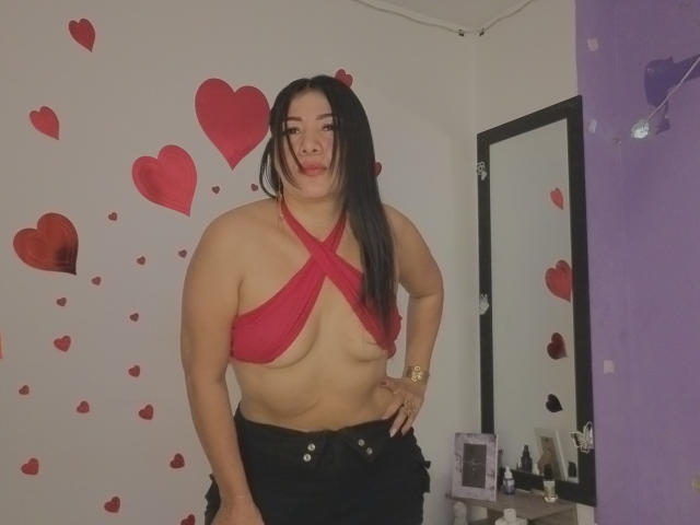 LuanaRose está em um quarto com adesivos de corações vermelhos, vestindo um top de biquíni vermelho com alças cruzadas, expondo seus seios grandes. calças pretas, pulseira dourada. Espelho, itens de maquiagem ao fundo. Incrivelmente sexy..