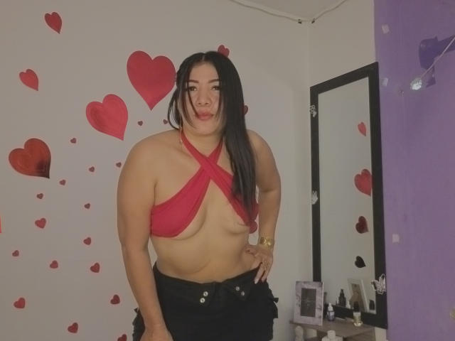 LuanaRose si trova in una stanza con adesivi a forma di cuore rosso sulle pareti. Indossa una maglia rossa a righe incrociate che mette in risalto il suo seno generoso, e dei pantaloni neri. La sua mano è appoggiata sul fianco.