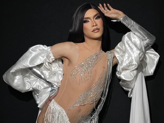 Agatha Rodriguez, di origine asiatica, con lunghi capelli neri, indossa un abito trasparente ricoperto di paillettes argentate, con maniche ampie e metallizzate, e ha una mano primo piano, sfondo nero, posa sicura, sexy, alta moda, glamour, ricami dettagliati, tessuto scintillante.