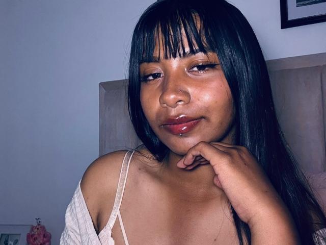 Close-up of NahomyNoir with black straight hair, bangs, and glossy red lips. She's wearing a white off-shoulder top, resting her chin on her hand, looking slightly to the side. Her skin is dark and smooth. She has a subtle, seductive expression.