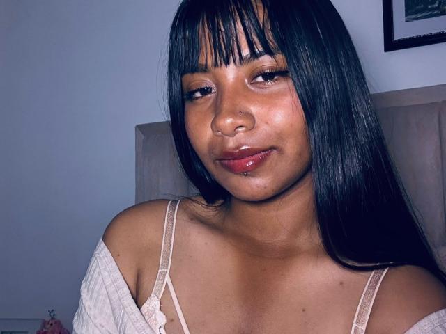 Nahaufnahme von NahomyNoir, einer kurvigen Frau mit dunkler Haut und langen schwarzen Haaren, die ein weißes Top mit einem Schulterauschnitt trägt und in die Ferne blickt. verführerisch mit glänzenden, roten Lippen, ihre Brust leicht entblößt.