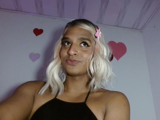 Fotografie von IdiiaLatina, einer kurvigen Frau mit hellbrauner Haut und platinblondem Haar, die ein schwarzes Top mit Halter trägt. rosa Haarspangen und rosa Herzdekorationen an der Wand. Sie blickt verführerisch in die Kamera..