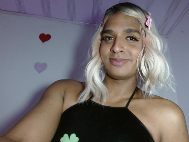 Photograph of IdiiaLatina, a Latina woman with platinum blonde hair, pink hairclip, and black halter top, smiling at the camera. Background has purple and red heart stickers. Sexual tension from confident gaze.