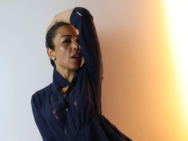 BrendaLive, dark-haired woman with slicked-back hair, wearing a black shirt, hand raised behind head, seductive expression, smooth skin, orange and blue gradient background, sexual tension, confident pose.