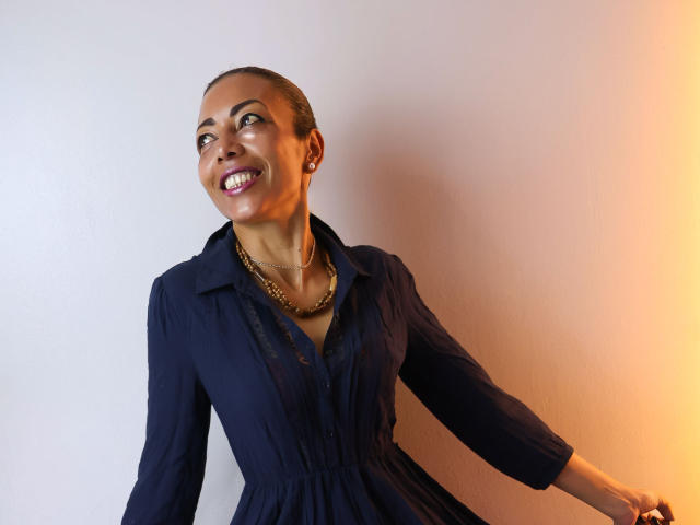 BrendaLive with dark skin, short hair, and a broad smile, wearing a black button-up shirt and gold chain, leaning against a white and orange wall, exuding confident sexual allure.