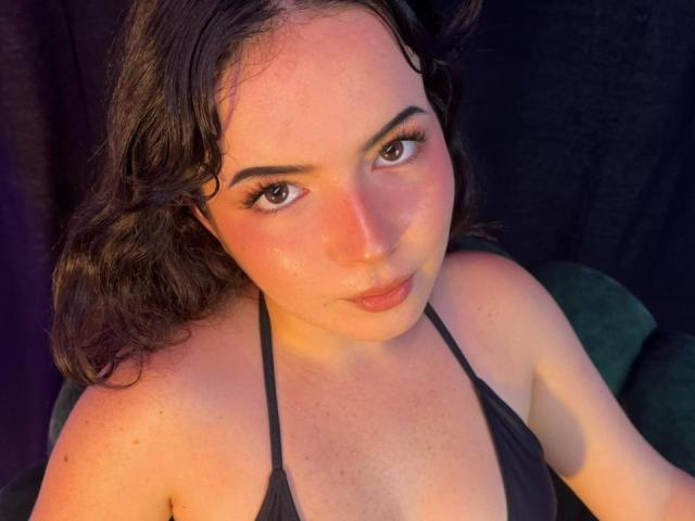 Close-up photograph of AliinaFontaine, a curvy woman with fair skin and dark wavy hair, wearing a black halter top, gazing seductively at the camera with intense brown eyes, soft light highlighting her face.