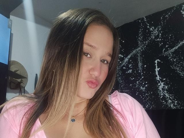Close-up of AlexaHotSeins, a young woman with long brown hair, pink lips, and a pink top. She's looking at the camera with a slightly pouty expression, creating sexual tension.
