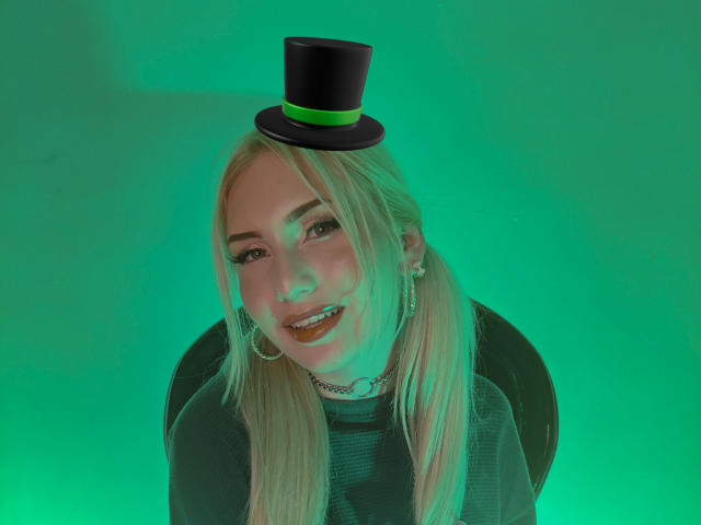 MariyLu, een blonde vrouw met steil haar, draagt een zwarte hoge hoed en een zwarte top, die groen licht uitstraalt. Met een glimlach draagt ze oorringen en een ketting. Ongelooflijk aantrekkelijk..